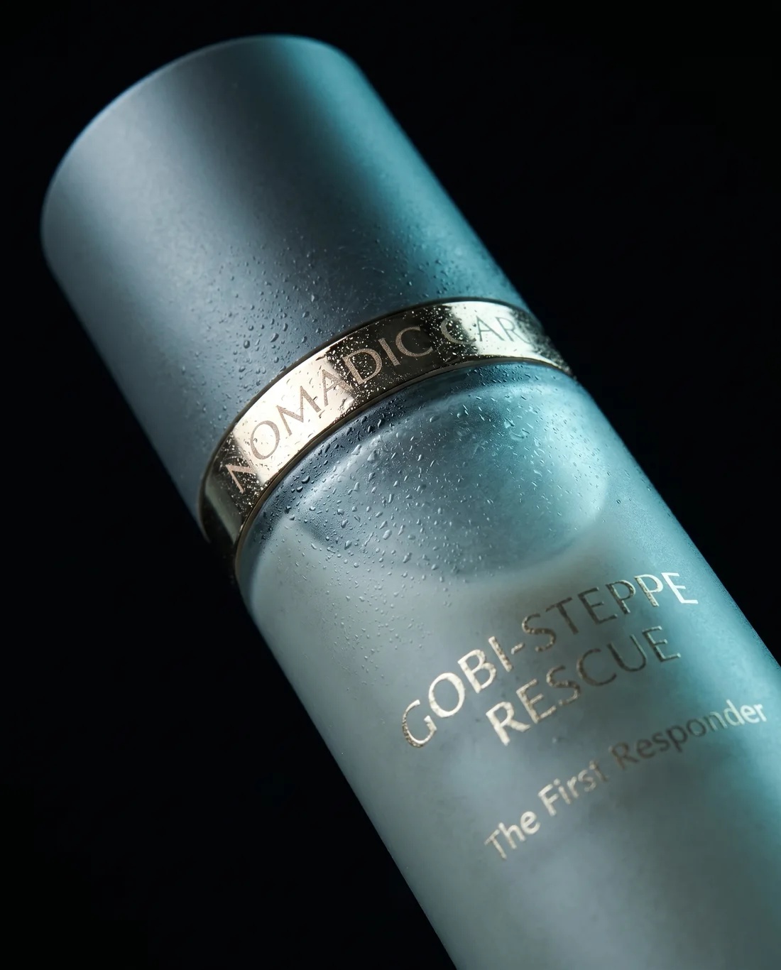 Macro detail of the Gobi-Steppe Rescue bottle: the gold Nomadic Care band and engraved label, water beads on frosted glass